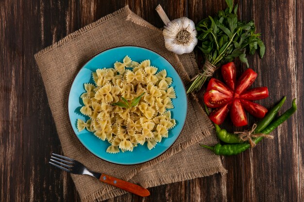 Top view of boiled pasta on a blue plate on a beige napkin with a fork tomatoes garlic and chili peppers on a wooden surface