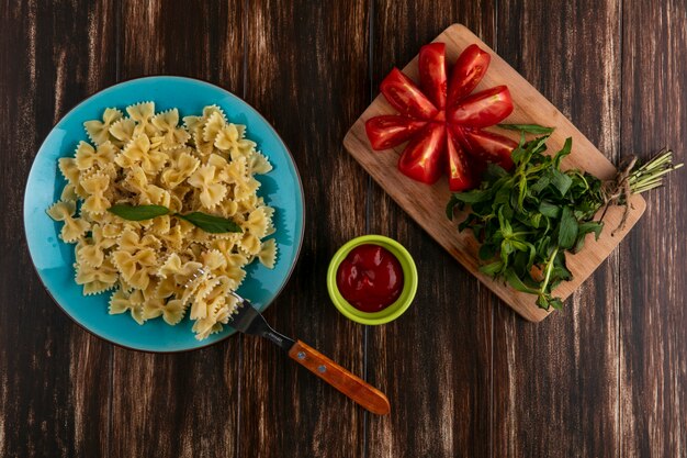 Top view of boiled pasta on a blue plate with a fork tomatoes and a bunch of mint on a cutting board with ketchup on a wooden surface