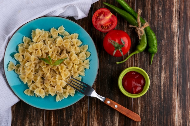 Top view of boiled pasta on a blue plate with a fork tomatoes ketchup and chili peppers on a wooden surface