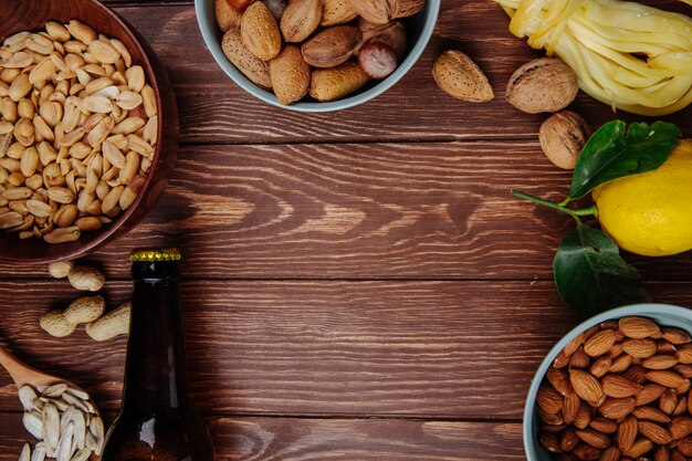 Top view of a bottle of beer with mix of salty snacks peanuts almond  string cheese with lemon on rustic wood with copy space
