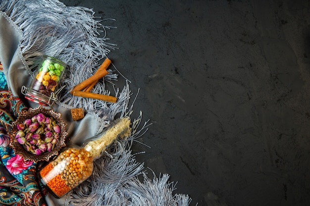 Top view of a bottle with raw corn seeds and beans dried rose buds and cinnamon stick on a shawl with tassel with copy space on black