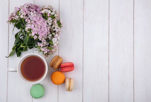 Top view of bouquet of flowers with a cup of tea and colored macarons on a white surface