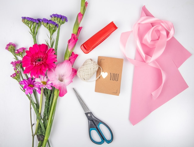 白い背景の上のスターチスとピンク色のガーベラとグラジオラスの花とピンクのリボンはさみと小さなポストカードと赤いホッチキスの花束のトップビュー