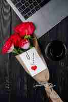 Free photo top view a bouquet of red roses in craft paper with attached postcard lying near a laptop with a paper cup of coffee on dark wooden background