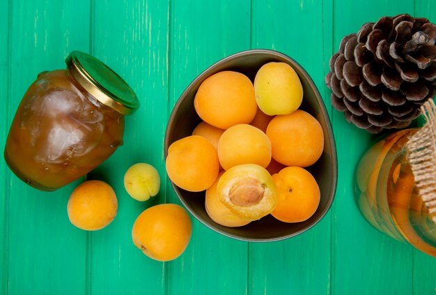 Top view of bowl of apricots and jar of peach jam with compote and pinecone on green background