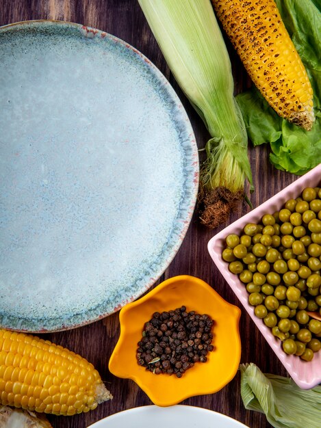 Free photo top view of bowl of black pepper with empty plate corns green peas on wooden surface