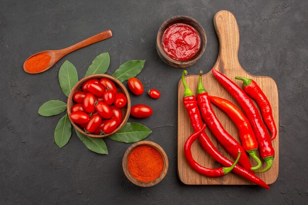 Top view a bowl of cherry tomatoes bay leaves hot red peppers on the chopping board a wooden spoon and bowls of ketchup and hot pepper powder on the black table