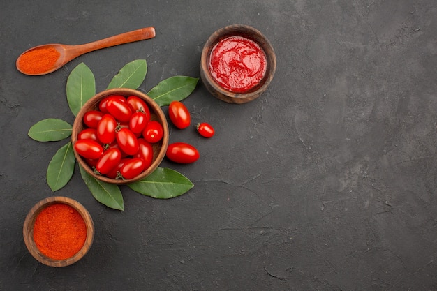 Top view a bowl of cherry tomatoes bay leaves a wooden spoon and bowls of ketchup and hot pepper powder on the black table