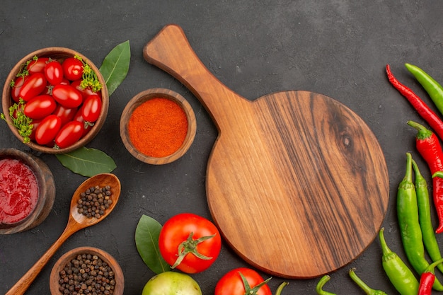 Top view a bowl of cherry tomatoes hot red and green peppers and tomatos bay leaves bowls of ketchup red pepper powder and black pepper and a chopping board on ground