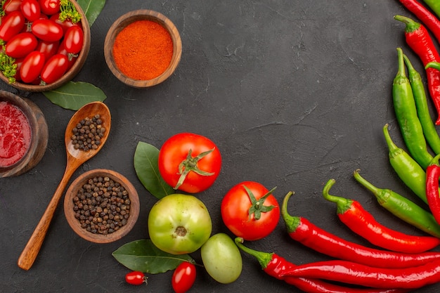 Top view a bowl of cherry tomatoes hot red and green peppers and tomatos bay leaves bowls of ketchup red pepper powder and black pepper and a spoon on black ground