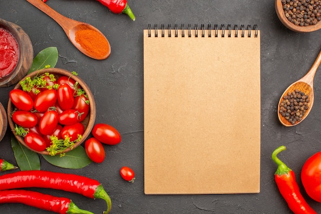 Top view a bowl of cherry tomatoes hot red peppers black pepper in a wooden spoon bowls of ketchup and black pepper and a notebook on black ground