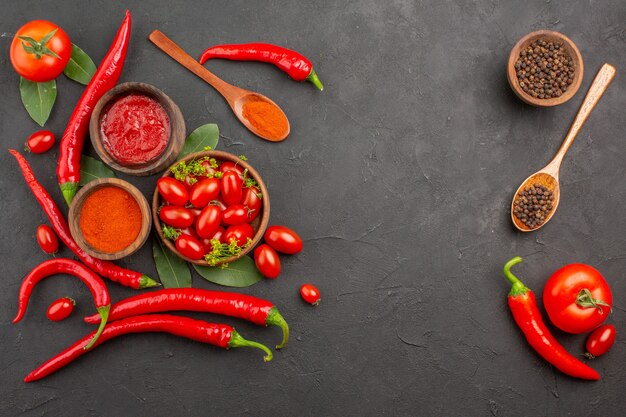 Top view a bowl of cherry tomatoes hot red peppers black pepper in a wooden spoon bowls of ketchup black pepper and red pepper powder on black ground