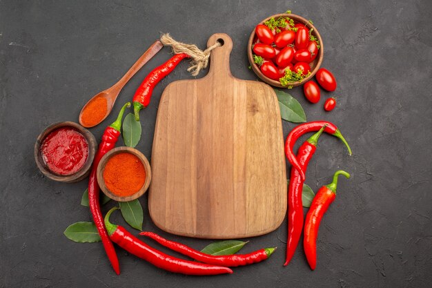 Top view a bowl of cherry tomatoes hot red peppers a chopping board a wooden spoon bay leaves and bowls of ketchup and hot pepper powder on the black table