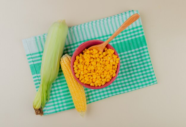 Top view of bowl of corn seeds with spoon and corn cobs on cloth and white surface