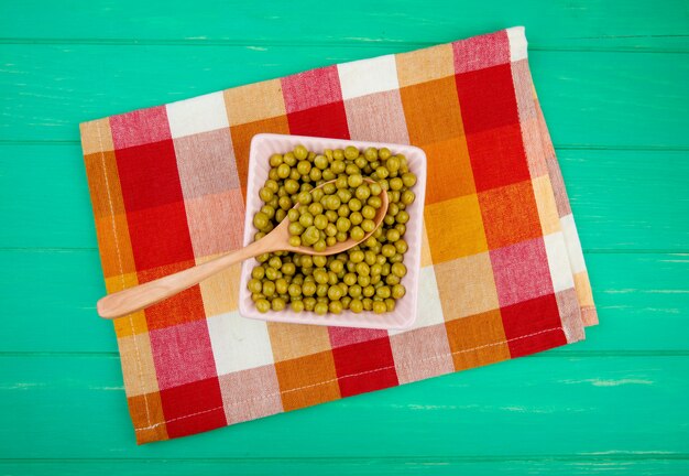 Top view of bowl of green pea with wooden spoon on cloth and green surface