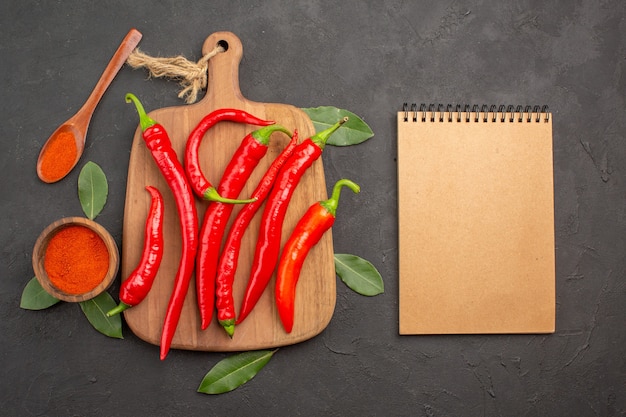 Top view a bowl of hot pepper powder red peppers on the chopping board bay leaves a wooden spoon and a notebook on the black table