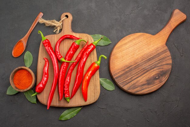 Top view a bowl of hot pepper powder red peppers on the chopping board bay leaves a wooden spoon and an oval chopping board on the black table