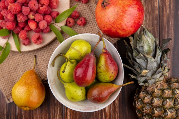 Top view of bowl of peach and raspberries on cutting board with pomegranate and peach on sackcloth and pineapple on wood