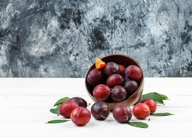 Top view a bowl of plums and strawberries on wicker placemat on white wooden board and dark blue marble surface.