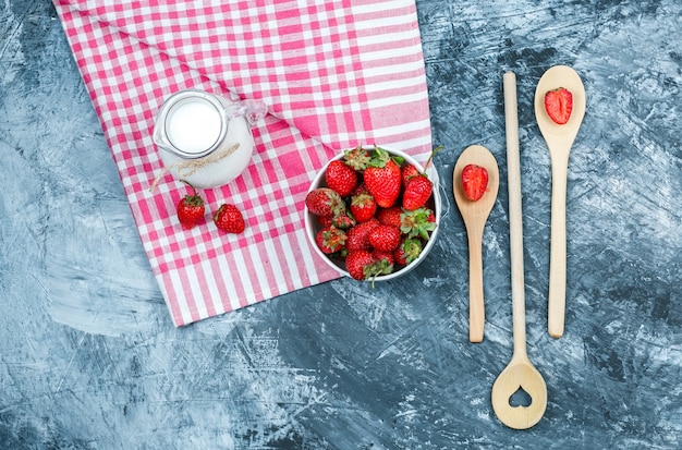 Top view a bowl of strawberries and a jug of milk on red gingham towel with wooden spoons on dark blue marble surface. horizontal