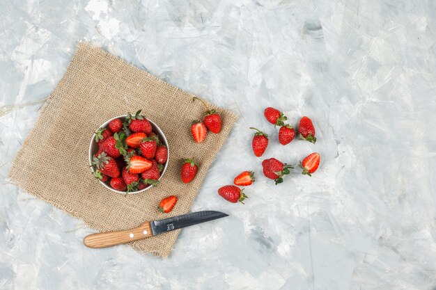 Top view a bowl of strawberries on a piece of sack with knife on white marble surface.