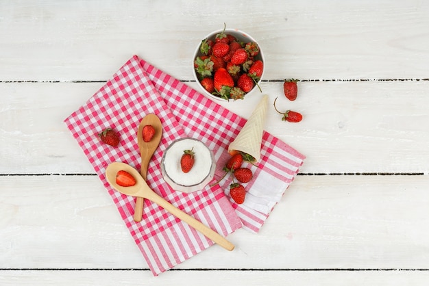 Top view a bowl of strawberries  on red gingham towel with wooden spoons,a cone of strawberries and a bowl of yogurt on white wooden board surface. horizontal