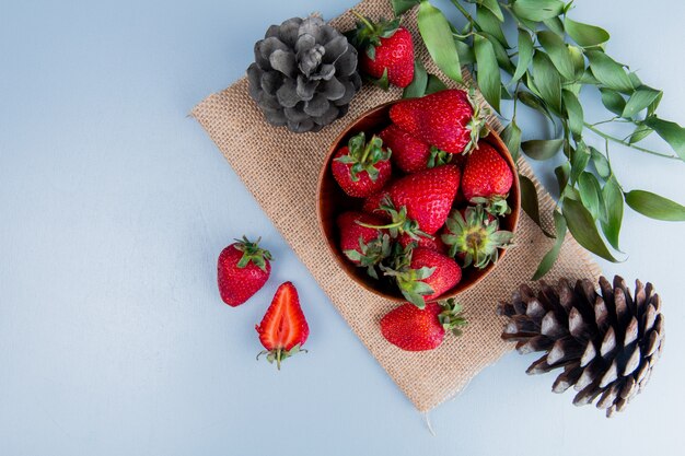 Top view of bowl of strawberries with pinecones on sackcloth on white surface decorated with leaves with copy space