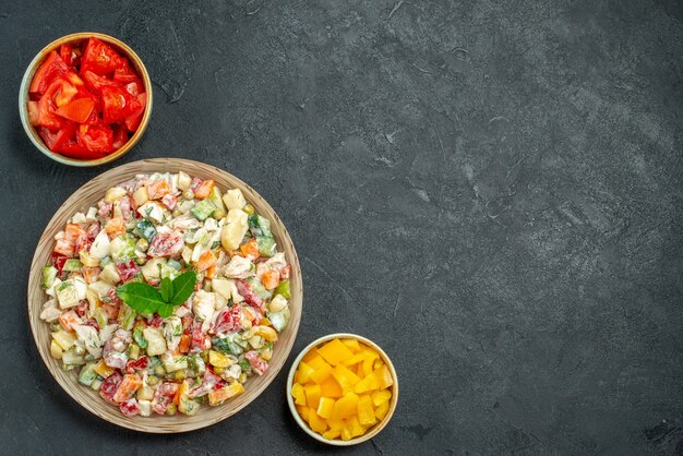 Top view of bowl of vegetable salad on left bottom side with bowls of vegetables on side on dark green-grey background