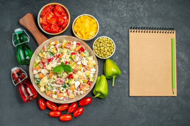 Top view of bowl of vegetable salad on plate stand with vegetables and oil and vinegar bottles and notepad on side and place for your text on dark grey background