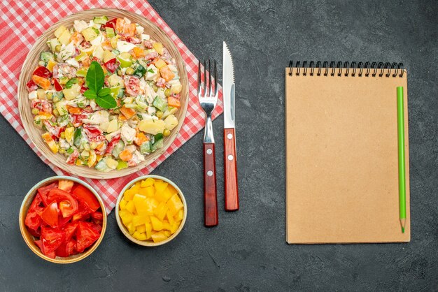 Top view of bowl of vegetable salad on red napkin with veggies cutleries and notepad on side on dark table