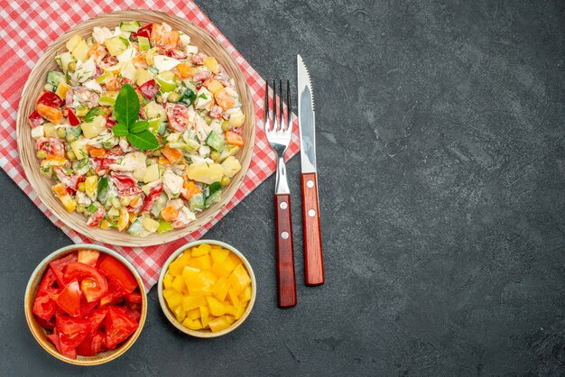 側面に野菜とカトラリーがあり、暗いテーブルにテキストのための自由な場所がある赤いナプキンの野菜サラダのボウルの上面図