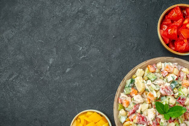 Top view of bowl of vegetable salad on right bottom side with bowls of vegetables on side on dark green-grey background