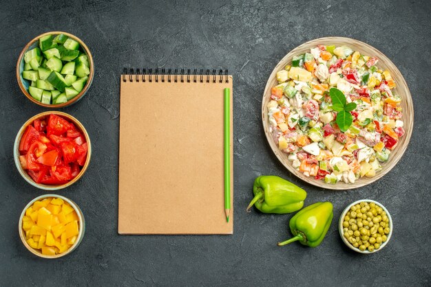 Top view of bowl of vegetable salad with bowls of vegetables notepad and bell peppers on side on dark green table