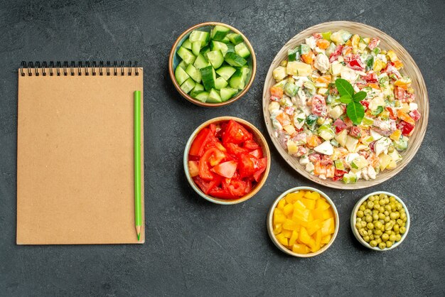 Top view of bowl of vegetable salad with bowls of vegetables and notepad with pen on it on side on dark green background