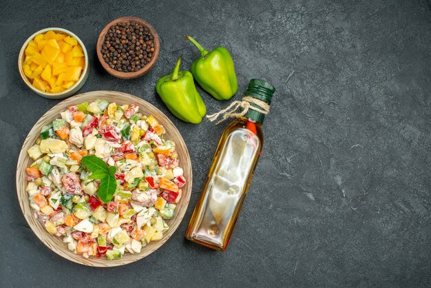 Вид сверху миски овощного салата с мисками овощей и бутылки перечного масла и болгарского перца сбоку на темном фоне
