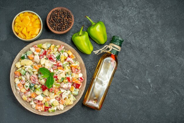 Top view of bowl of vegetable salad with bowls of vegetables and pepper oil bottle and bell peppers on side on grey background