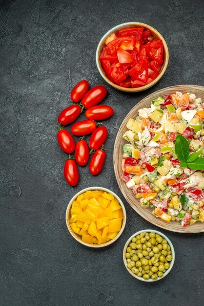 Top view of bowl of vegetable salad with vegetables on side on dark grey background