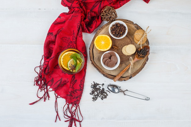 クッキーとクローブのボウル、ハーブティー、赤いスカーフ、白い表面の茶漉しと木の板に柑橘系の果物の上面図