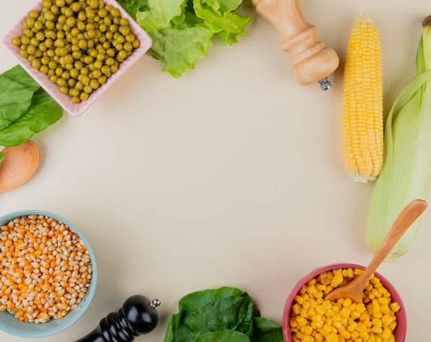 Top view of bowls of dried and cooked corn seeds green peas lettuce spinach corn cobs on white surface with copy space