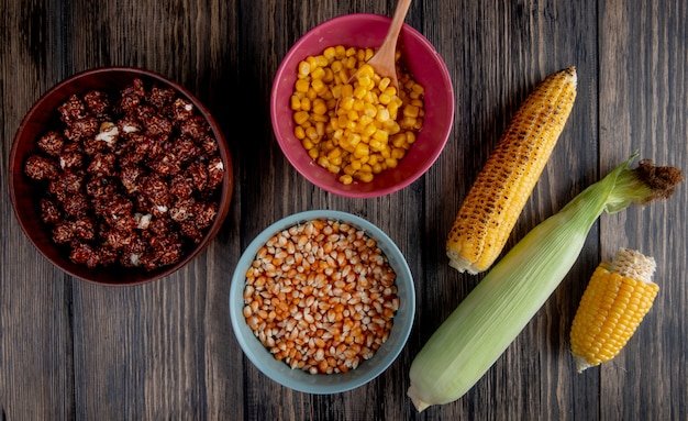 Top view of bowls full of chocolate popcorn with cooked and dried corn seeds and corns on wooden surface