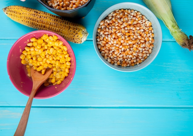 Вид сверху чаши, полные вареных и сушеных семян кукурузы с початками кукурузы на синей поверхности