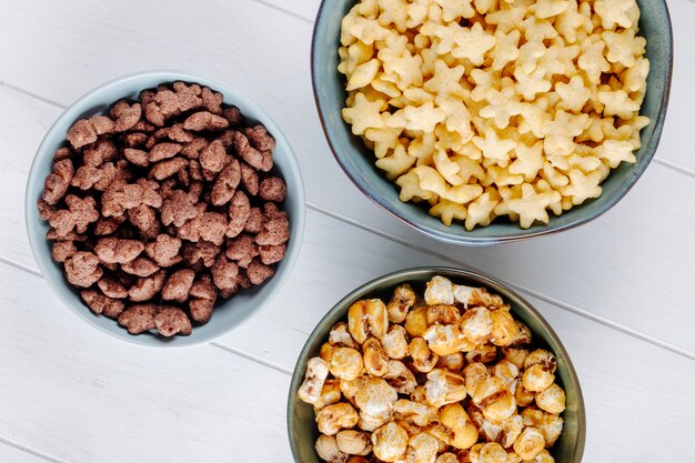 Top view of bowls of various cereals and sweet caramel popcorn on white wooden background