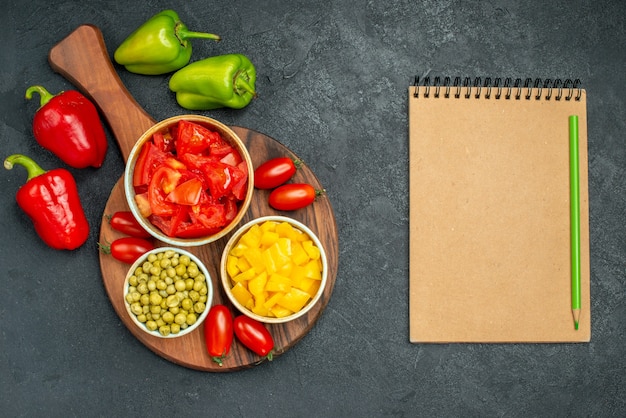 Top view of bowls of vegetables on plate stand with vegetables and notepadon side on dark grey background