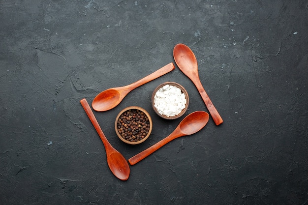 Free photo top view bowls with sea salt and black pepper around wooden spoons on dark table with copy place