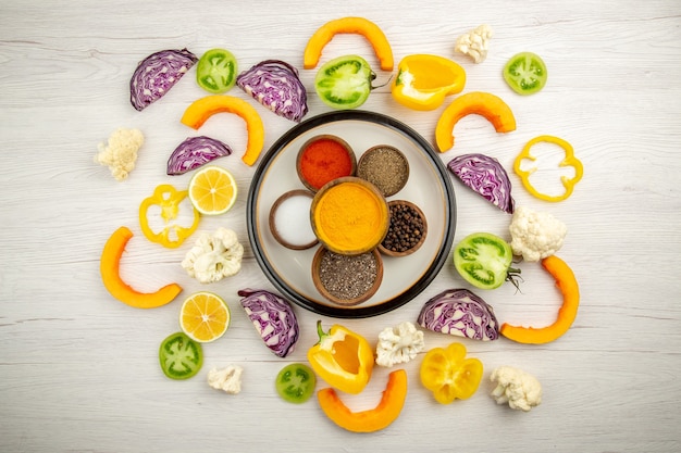 Бесплатное фото Вид сверху миски со специями на круглой тарелке, куркума, соль, черный перец, красный перец, нарезанные овощи на белой поверхности