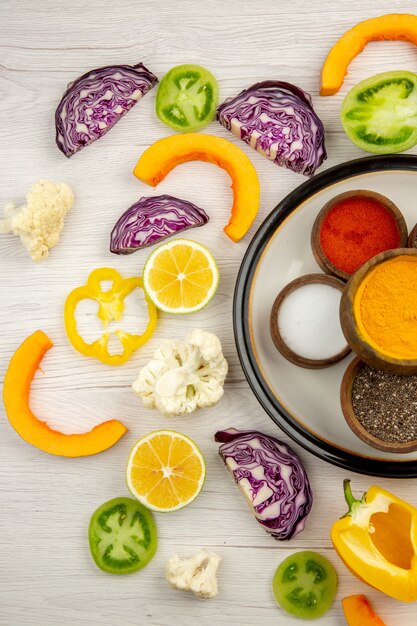 Top view bowls with spices on round plate cut vegetables on white surface