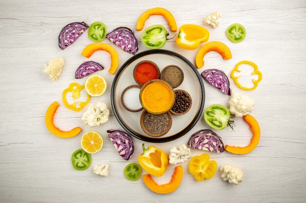 Top view bowls with spices on round plate turmeric salt black pepper red pepper chopped vegetables on white surface