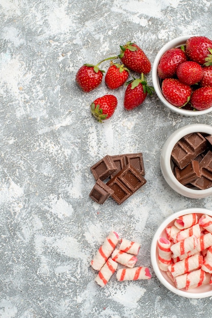 Top view bowls with strawberries chocolates candies and some strawberries chocolates candies at the right side of the grey-white table