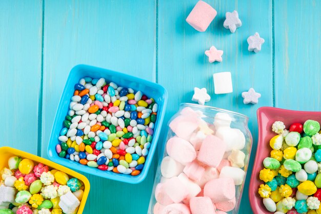 Top view of bowls with various colorful candies and marshmallow scattered from a glass jar on blue background