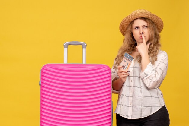 Top view of brainstorming young lady wearing hat holding ticket and standing near her pink bag